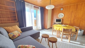 Een gezellige woonkamer met een grijze bank, een houten eettafel met een gele tafelkleed, en een televisie aan de muur.