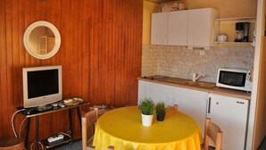 Een compacte keuken met een magnetron, spoelbak en witte kastjes. Er is een klein zitehoekje met een gele tafel en twee potplanten.