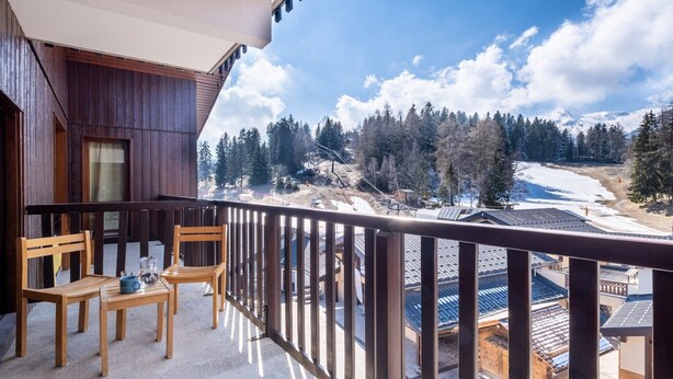 Un balcon avec des chaises et une table en bois, donnant sur un paysage enneigé parsemé d’arbres et de bâtiments.