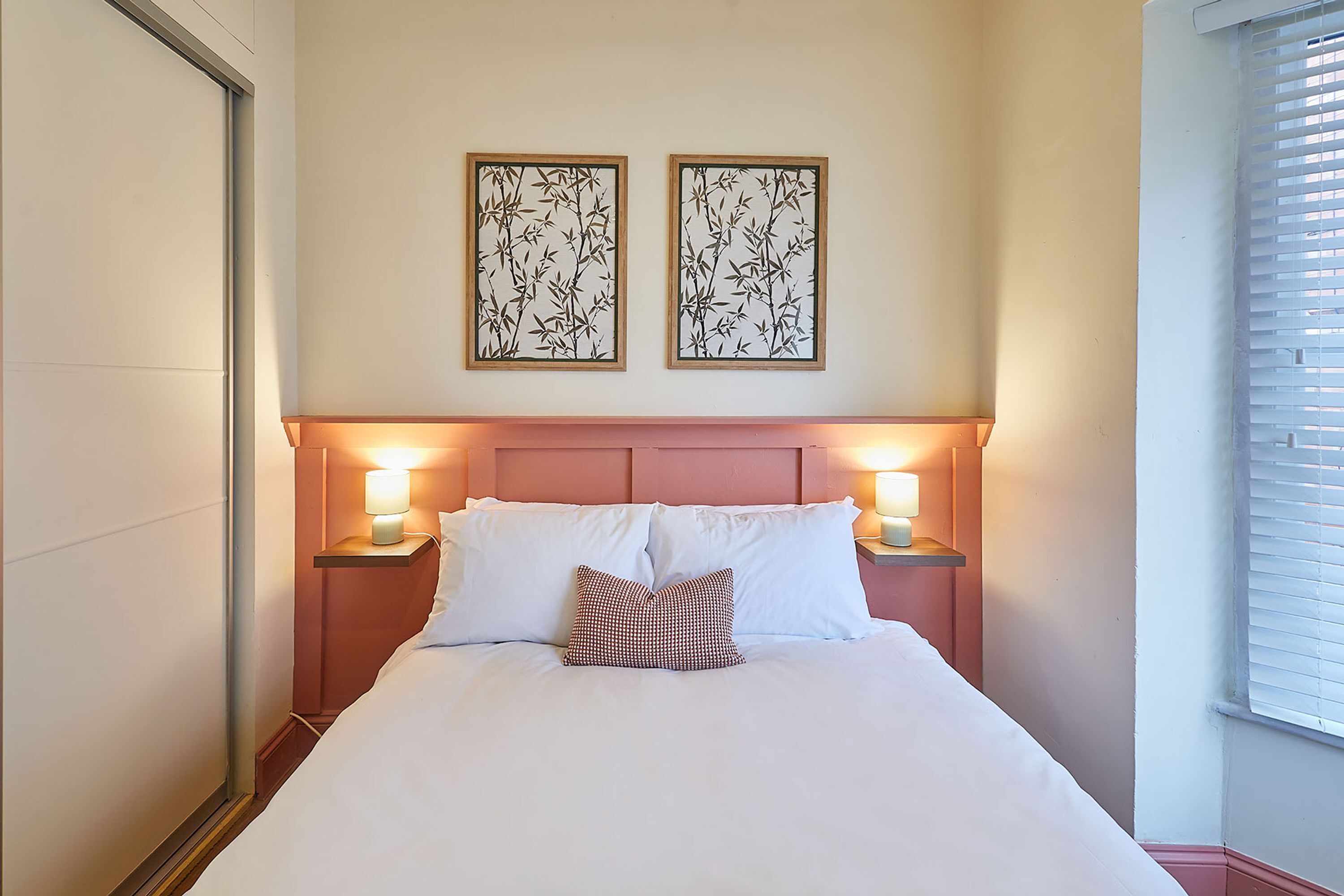 A neatly made bed with white linens and a patterned pillow, flanked by two bedside tables with lamps, in a room with a simple, clean design and two framed wall art pieces above the bed.