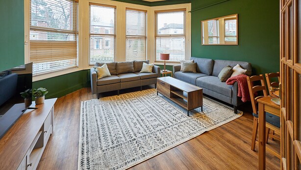A living room with a sectional sofa, a coffee table, and a patterned rug. There are large windows with blinds, a small table with a lamp, and a chair. - Host Stay Apartment 1 St Stephens Court (Worcester)
