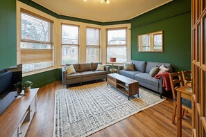 A living room with a sectional sofa, a coffee table, and a patterned rug. There are large windows with blinds, a small table with a lamp, and a chair.