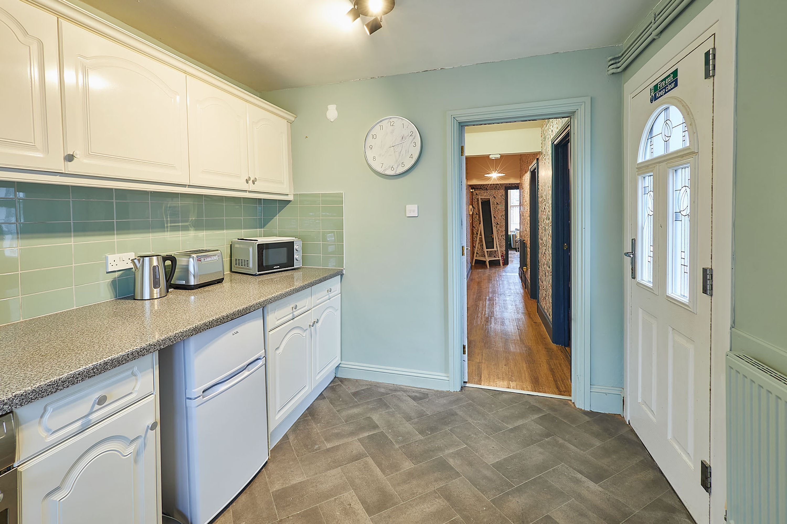 A kitchen with white cabinets, a microwave, and a toaster. There is a clock on the wall and a door leading to another room.