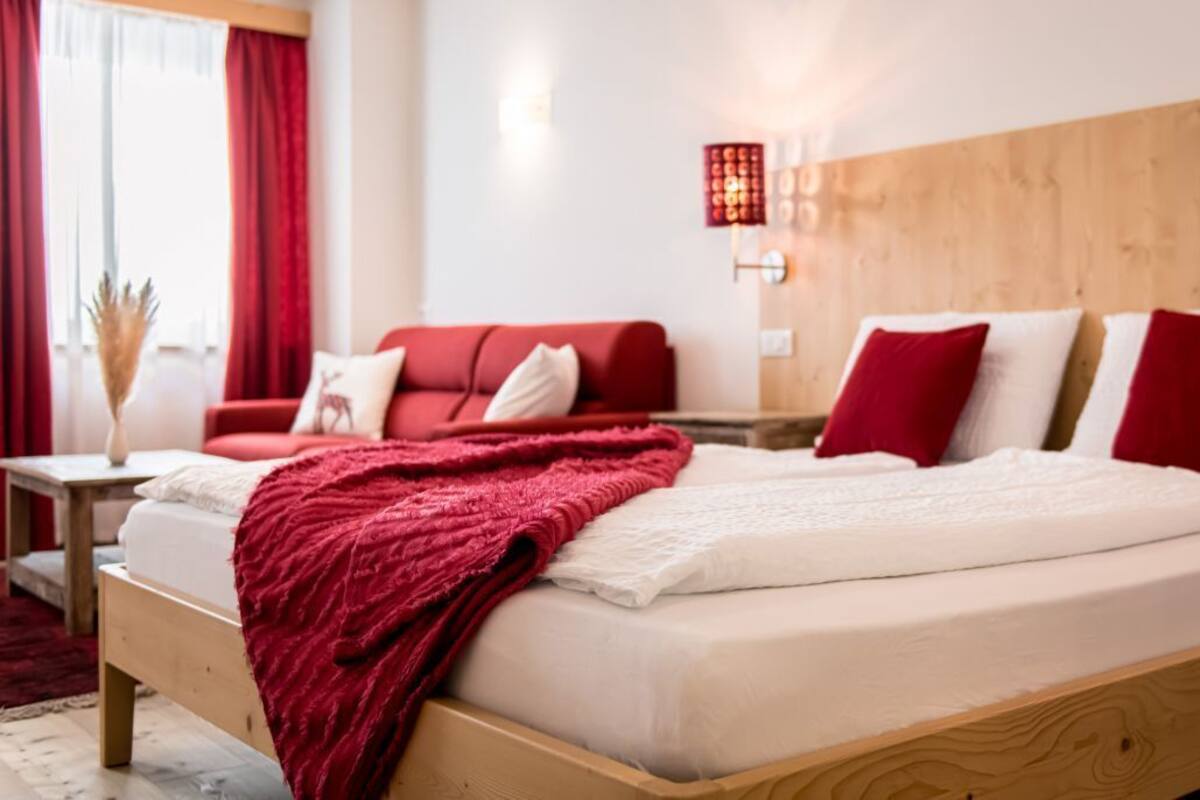 Une chambre d’hôtel avec un lit en bois, une literie rouge et blanche, un canapé rouge et une petite table sur laquelle est posé un vase.
