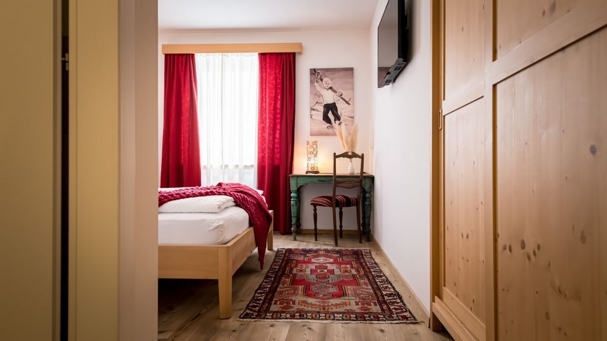 A bedroom with a bed, a wooden wardrobe, a small table with a lamp, and a red curtain.