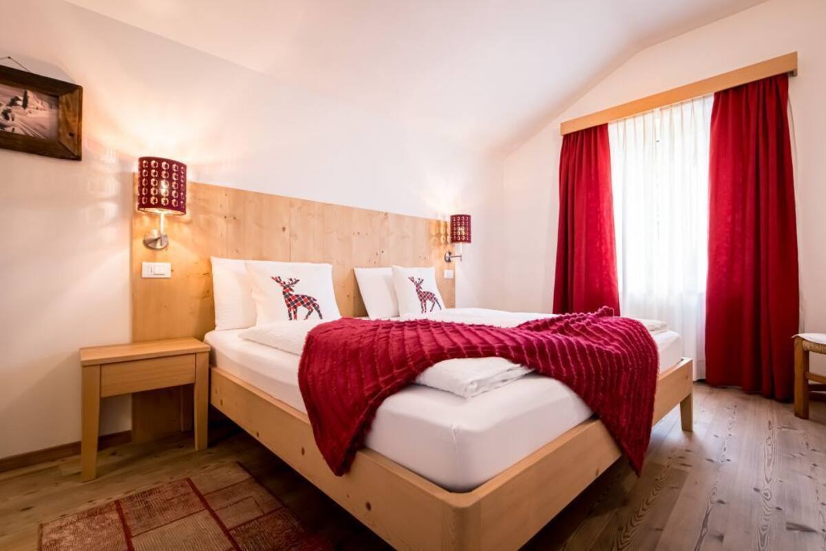 Une chambre à coucher avec une tête de lit en bois, un lit recouvert de draps blancs et d’une couverture rouge, deux lampes de chevet, une table de chevet en bois, un rideau rouge et une chaise en bois.