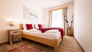 A bedroom with a wooden bed, white bedding, red pillows, a nightstand with a lamp, a window with curtains, and a decorative vase. - Am Schloss Guest House (Brunico)