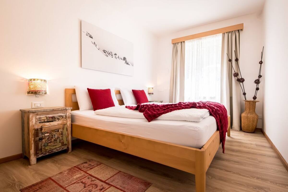 Une chambre à coucher avec un lit en bois, du linge de lit blanc, des coussins rouges, une table de chevet avec une lampe, une fenêtre avec des rideaux et un vase décoratif.
