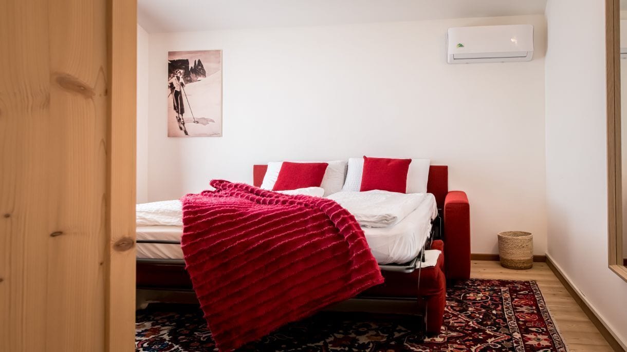 A bedroom with a bed, red and white bedding, a red throw blanket, a red chair, a small rug, and a wall-mounted air conditioner.