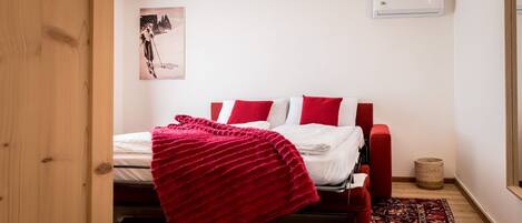 A bedroom with a bed, red and white bedding, a red throw blanket, a red chair, a small rug, and a wall-mounted air conditioner.