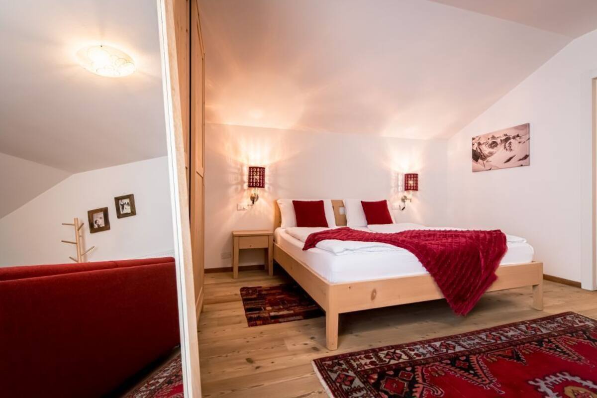 Une chambre avec un lit en bois, des oreillers rouges, un plaid rouge et un tapis à motifs. Il y a deux lampes fixées au mur et un luminaire au plafond.