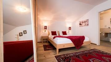 A bedroom with a wooden bed, red pillows, a red throw blanket, and a patterned rug. There are two wall-mounted lights and a ceiling light.