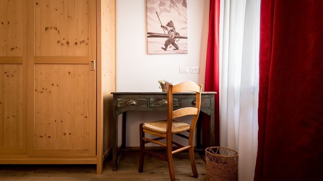 Una habitación con un armario de madera, un escritorio con silla, un cuadro en la pared, una cortina roja y una cesta de mimbre.