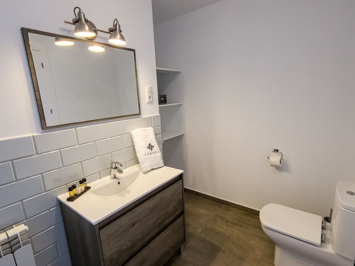 Un bagno moderno con un lavandino bianco, una cassettiera in legno scuro, un grande specchio illuminato e un water bianco.