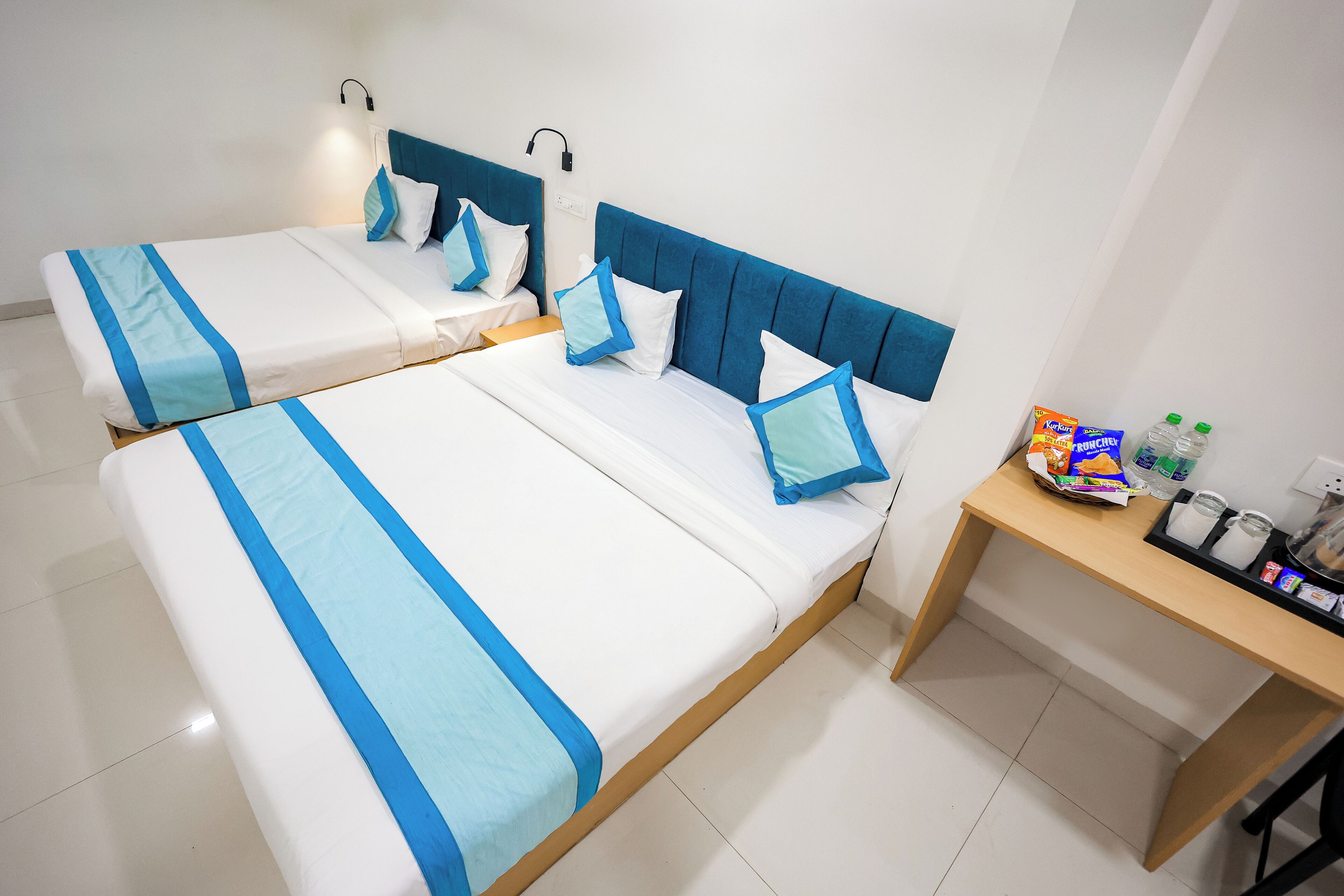 A hotel room with two beds, each with blue and white bedding, a wooden nightstand with toiletries, and a wall-mounted lamp.