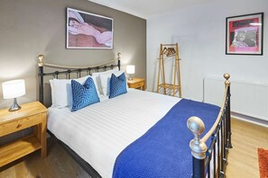 A neatly made bed with a blue blanket and patterned pillows, a wooden nightstand with a lamp, and a wooden hanger rack against the wall. - Host Stay Garden Flat (Brighton)