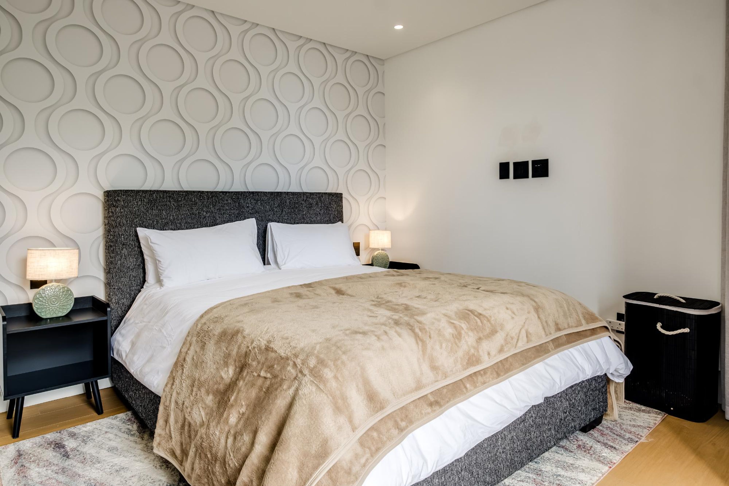A modern bedroom with a large bed, a patterned wall, and a nightstand with a lamp.