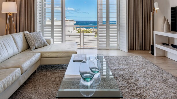 Un salon moderne avec une table basse en verre, un canapé beige et une vue sur l’océan grâce à de grandes fenêtres.