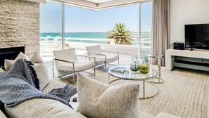 A modern living room with a large window overlooking the sea, a flat-screen TV, and a cozy seating area. - Sapphire Tide Penthouse w Sea View Private Pool (Cape Town)