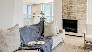 A modern living room with a stone fireplace, a sofa with a patterned throw, and a view of the kitchen and balcony. - Sapphire Tide Penthouse w Sea View Private Pool (Cape Town)