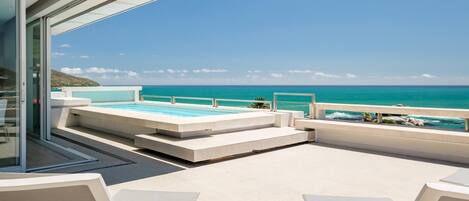 A modern outdoor pool area with a clear view of the ocean, a glass-enclosed room, and a seating area with tables and chairs.
