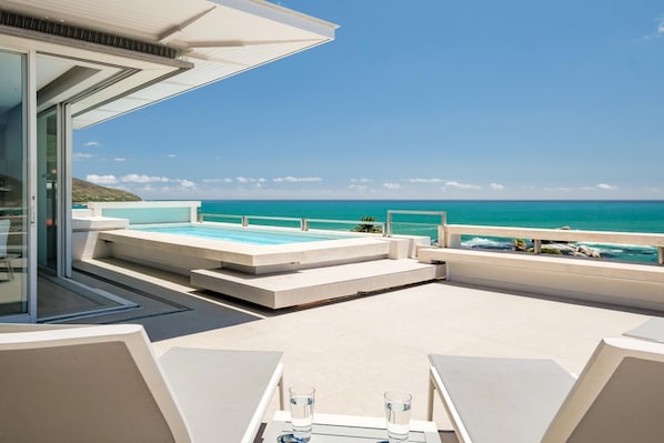 A modern outdoor pool area with a clear view of the ocean, a glass-enclosed room, and a seating area with tables and chairs.