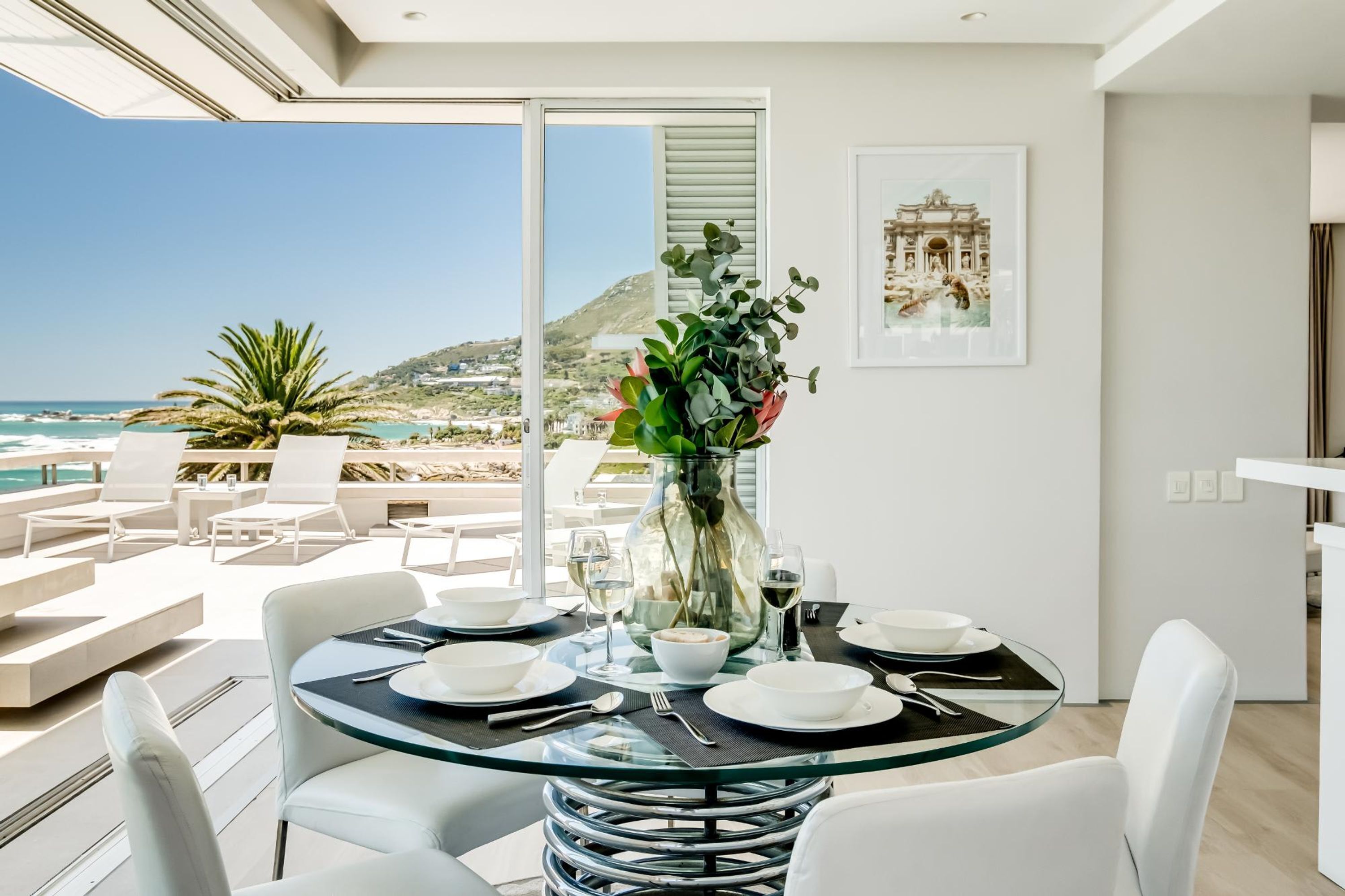 Un comedor moderno con una mesa de vidrio, sillas blancas y vista al océano.