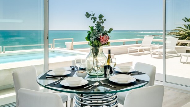 A dining area with a glass table and chairs, a vase with flowers, and a view of the ocean. - Sapphire Tide Penthouse w Sea View Private Pool (Cape Town)