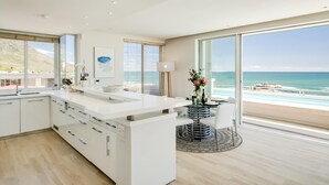 A modern kitchen with a large island, a dining area with a view of the beach, and a large window overlooking the ocean. - Sapphire Tide Penthouse w Sea View Private Pool (Cape Town)