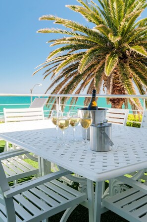 Een witte buitentafel met vier wijnglazen en een champagnefles, voor een achtergrond van een palmboom en een helderblauwe lucht.