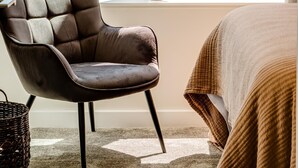 Quarto moderno com uma grande janela, uma poltrona acolchoada, uma pequena planta em vaso e uma cama com manta texturizada.