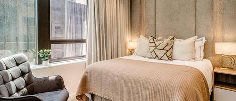 A modern hotel room with a large bed, a dark leather chair, a nightstand, and a window with curtains.