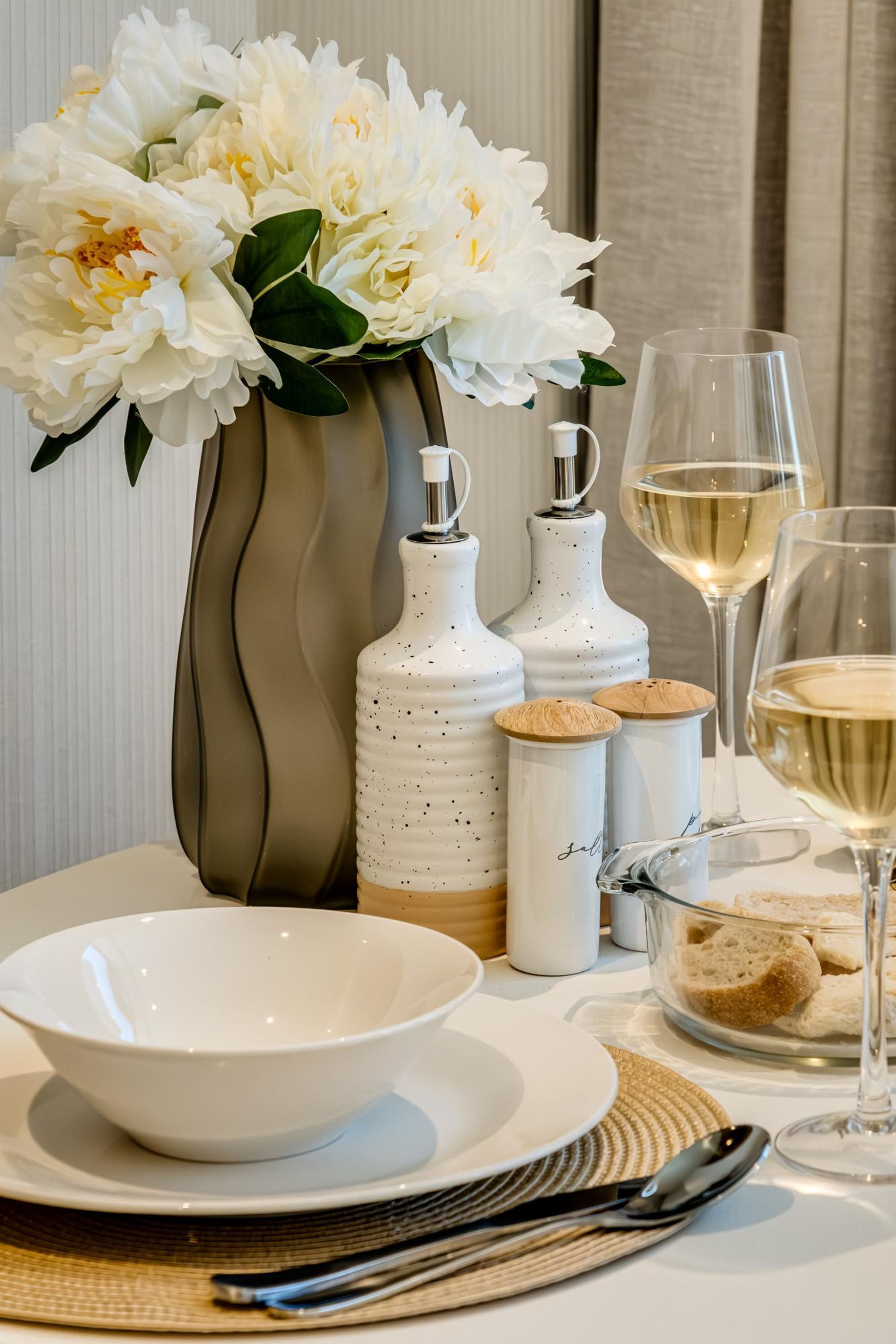 A table set with white flowers, a vase, two condiment shakers, a glass of white wine, bread, and a bowl.