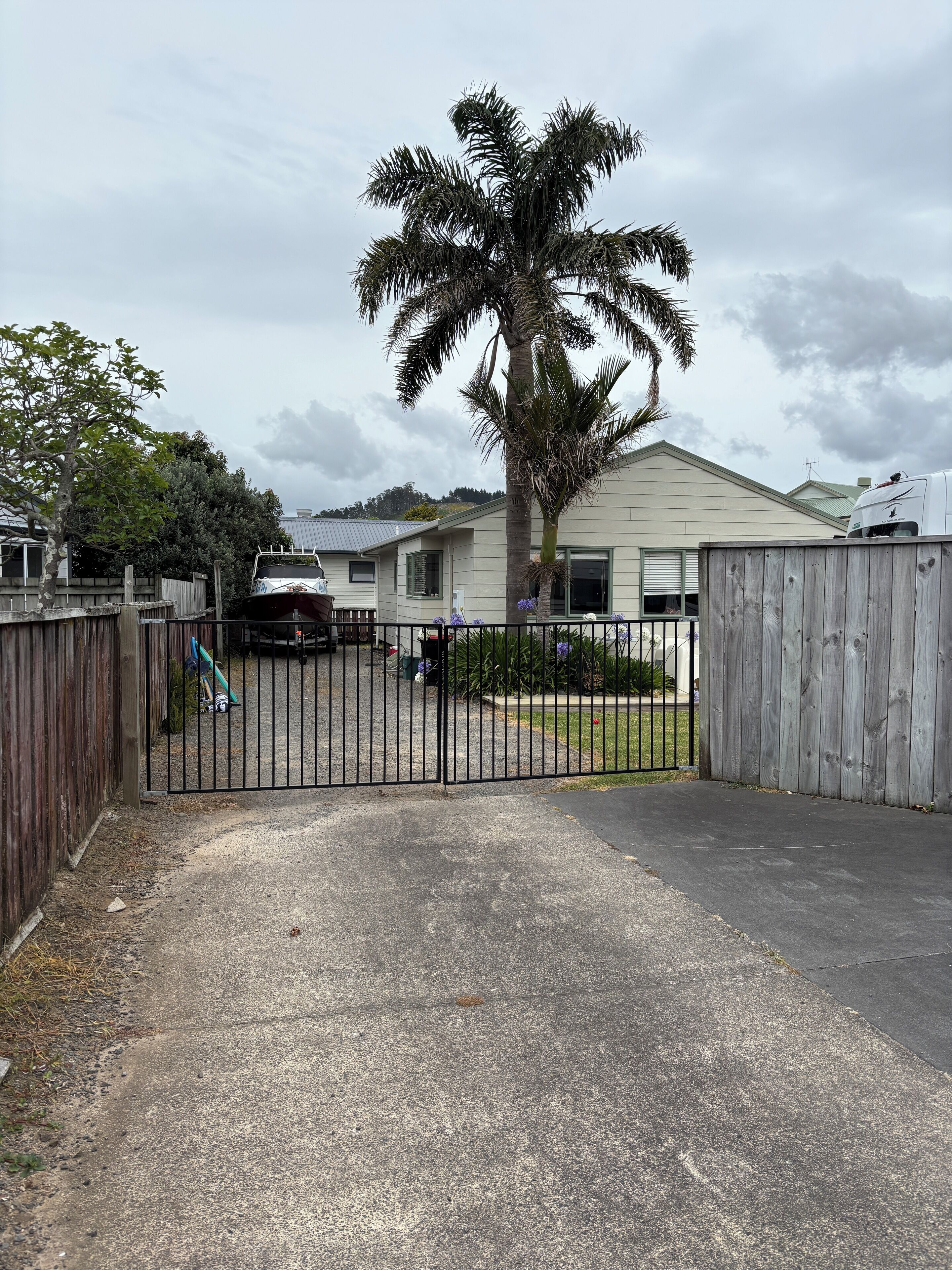 Une allée fermée par un portail mène à une maison ornée d’un palmier et devant laquelle est garé un bateau.