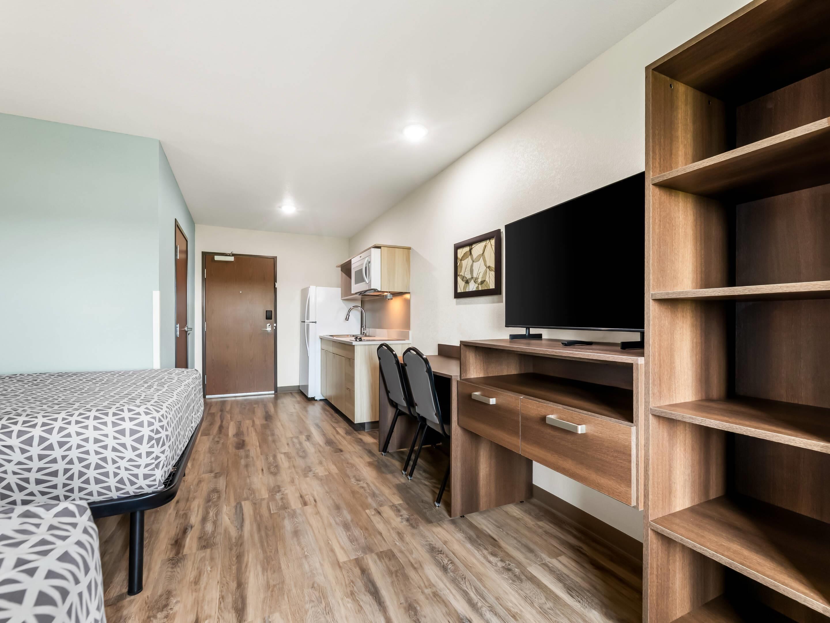 A compact kitchenette with a microwave, refrigerator, and a flat-screen TV on a wooden shelf.