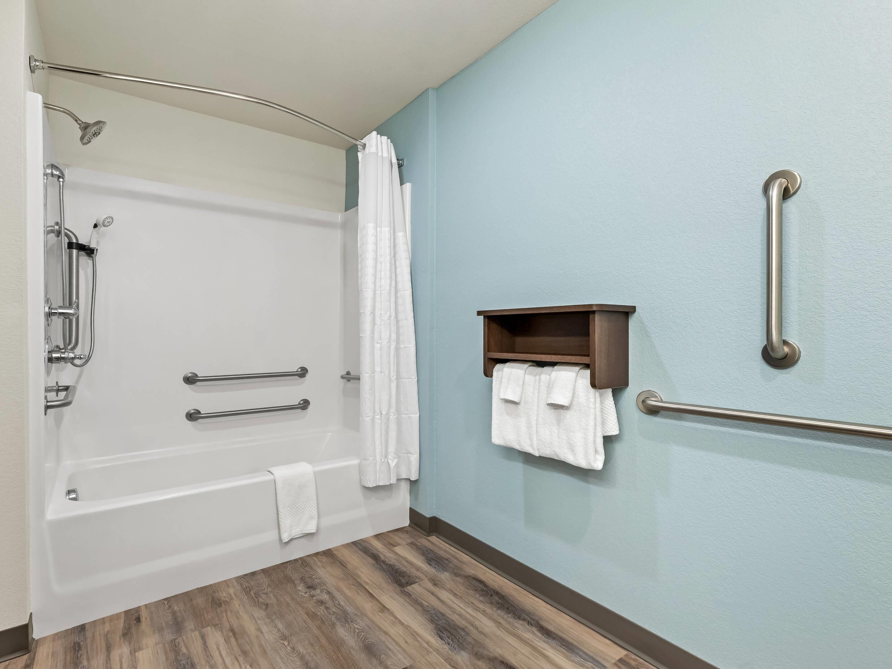 Un baño con bañera, barras de apoyo, un perchero y un estante de madera.