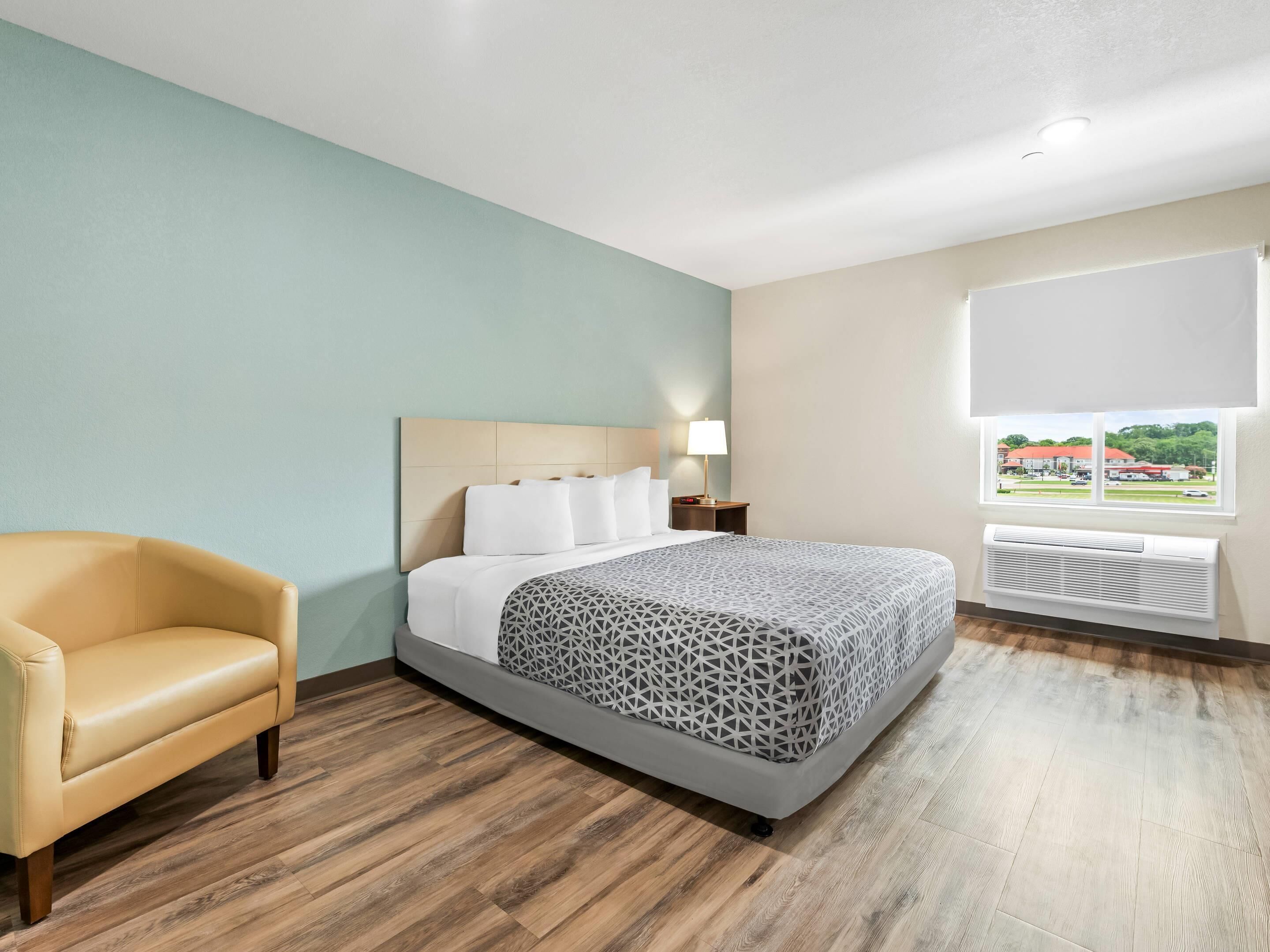 A hotel room with a bed, a chair, a nightstand, a lamp, and a window with a view of buildings.