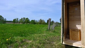 A wooden structure with an open door, a fenced area, and a grassy field with trees in the background. - Bee wagon with fireplace & sauna can be used all year round in the natural garden (Dargun)