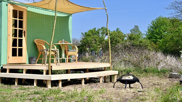 A green wooden structure with a porch, wicker chairs, and a barbecue grill. - Bee wagon with fireplace & sauna can be used all year round in the natural garden (Dargun)