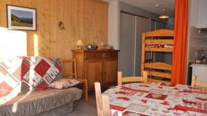 A cozy room with a wooden bunk bed, a sofa with a quilt, a table with a patterned tablecloth, and a kitchen area with a sink and cabinets.