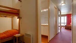A bunk bed with a red blanket, a small bedside table, and a radiator. A hallway leads to a bathroom with a sink and a window with red curtains. - Cozy Retreat with Ski Access (Les Deux Alpes)