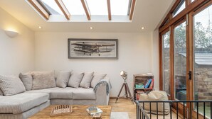 A living room with a sectional sofa, a wooden coffee table with chess pieces, a framed airplane picture, a tripod lamp, and a bookshelf. - Coach House - Lovely Home in Cheltenham (Cheltenham)