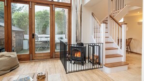 A cozy living room with a fireplace, a staircase, and a view of the garden through large glass doors. - Coach House - Lovely Home in Cheltenham (Cheltenham)