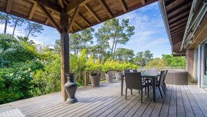 Una terraza de madera con mobiliario exterior, rodeada de exuberante vegetación y árboles altos.