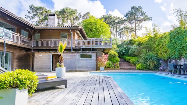 Una terraza de madera con piscina, rodeada de exuberante vegetación y una casa de dos pisos con balcón.