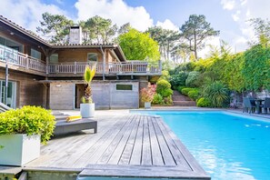 A wooden deck with a pool, surrounded by lush greenery and a two-story house with a balcony. - Villa d'Architecte piscine / dune & plage a pied (Pyla-sur-Mer)