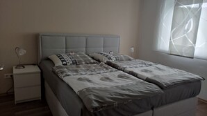 Cama doble con cabecera gris, dos mesitas de noche y una ventana con cortinas de gasa.