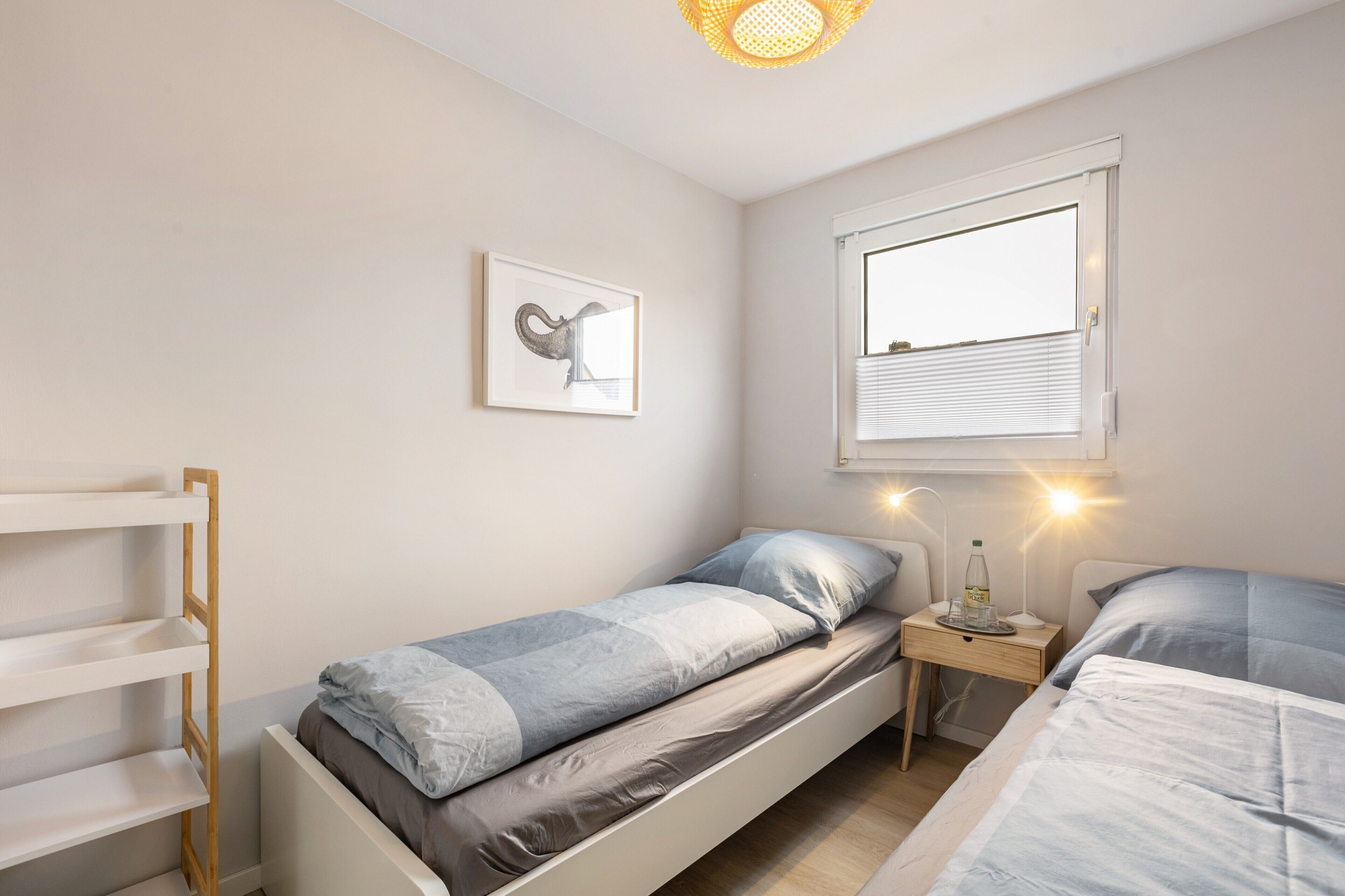 Une chambre avec deux lits, une table de chevet avec une lampe, une étagère fixée au mur et une fenêtre avec des stores.