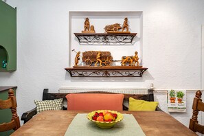 Uma mesa de jantar com uma fruteira, um travesseiro verde xadrez e um travesseiro amarelo. Há cadeiras de madeira e uma parede verde com uma janela.