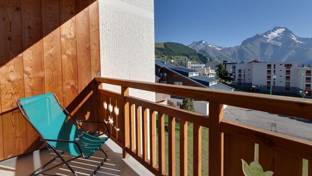 Ein Balkon mit Holzbalkon, einem türkisfarbenen Stuhl und Blick auf Gebäude und Berge im Hintergrund.